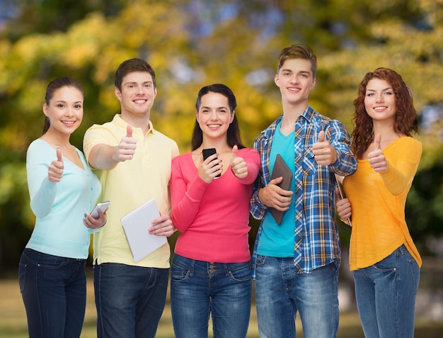 concepto de amistad, tecnología, gestos y personas - grupo de adolescentes sonrientes con teléfonos inteligentes y tabletas mostrando los pulgares hacia arriba sobre el fondo del parque