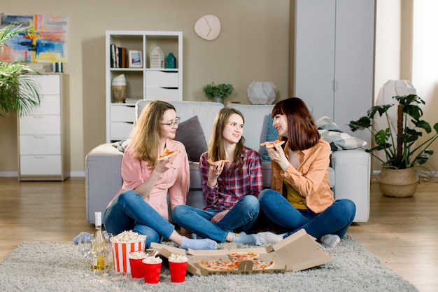 Concepto de amistad, vacaciones, comida rápida y celebración: amigas felices con bebidas y palomitas de maíz comiendo pizza en casa, sentados en el suelo en una habitación acogedora