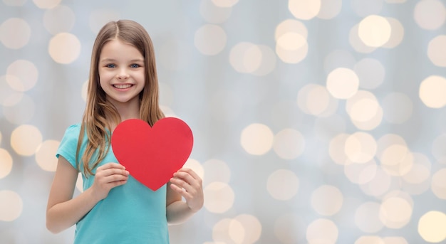 concepto de amor, caridad, vacaciones, niños y personas - niña sonriente con corazón rojo sobre fondo de luces