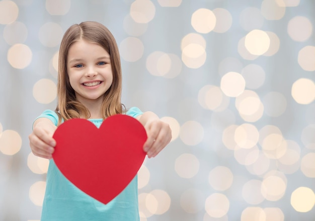 concepto de amor, caridad, vacaciones, niños y personas - niña sonriente con corazón rojo sobre fondo de luces