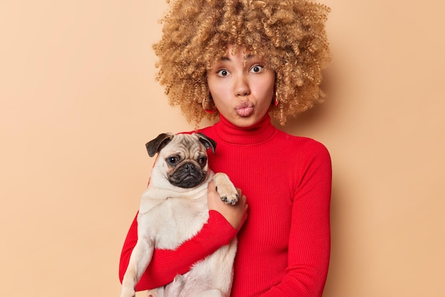 Concepto de amor y mascotas de la gente El afectuoso dueño del perro envía mwah a la cámara mantiene los labios plegados en poses con un cachorro que viste un cuello de tortuga rojo casual aislado sobre un fondo beige Amante de los animales Foto de estudio