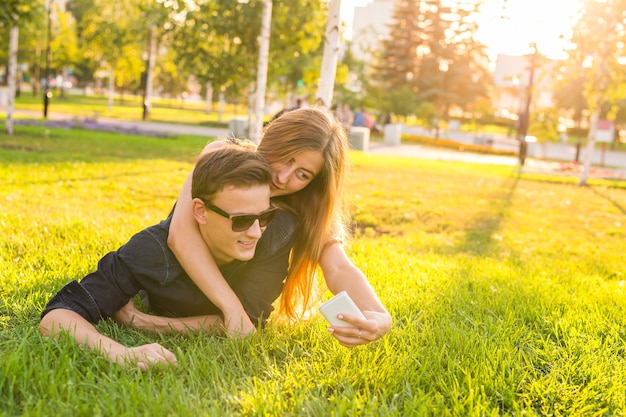 Concepto de amor y personas - feliz pareja adolescente tumbado en la hierba y tomando selfie en smartphone en verano.