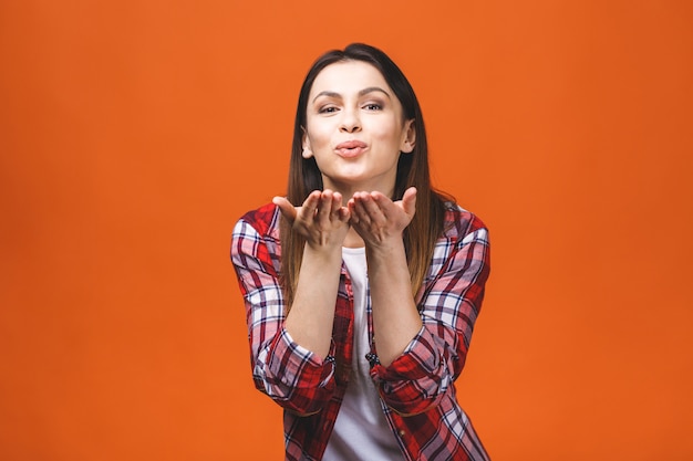 Concepto de amor Retrato de una joven morena posando soplando un beso, aislado sobre fondo naranja.