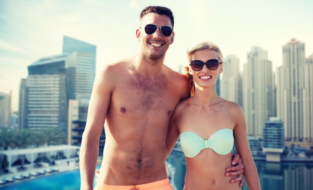 concepto de amor, viajes, turismo, verano y personas - pareja sonriente de vacaciones en traje de baño y gafas de sol abrazándose sobre el fondo frente al mar de la ciudad de dubai