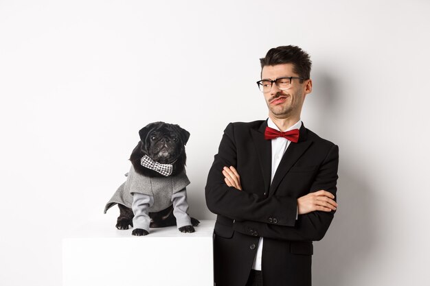 Concepto de animales, fiesta y celebración. Imagen de un joven divertido con traje y gafas, mirando escéptico al lindo pug disfrazado, de pie sobre blanco.