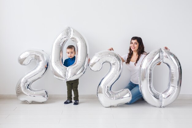 Concepto de año nuevo, celebración y vacaciones: madre e hijo sentados cerca del letrero 2020 hecho de globos plateados para año nuevo en sala blanca