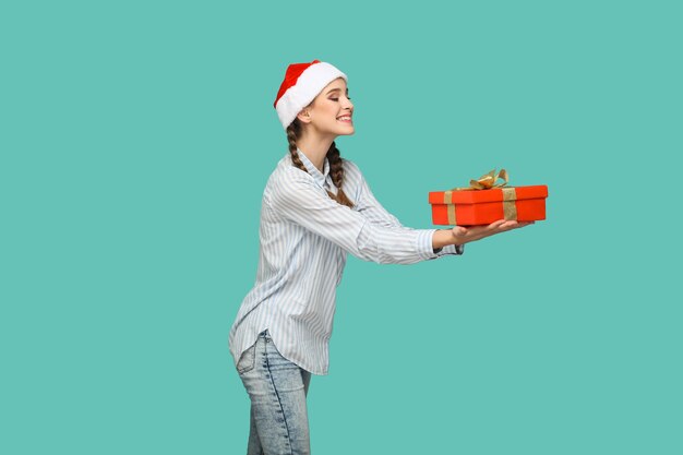 Concepto de año nuevo. Vista lateral del perfil de la hermosa chica en camisa azul claro a rayas en gorro rojo de Navidad de pie y dando caja de regalo roja con una gran sonrisa. interior, aislado sobre fondo verde.