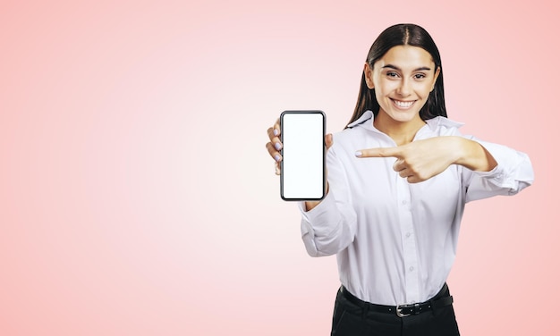Concepto de aplicación móvil con una chica feliz con camisa blanca que muestra un teléfono inteligente moderno con una pantalla blanca en blanco sobre fondo rosa claro abstracto con espacio de copia