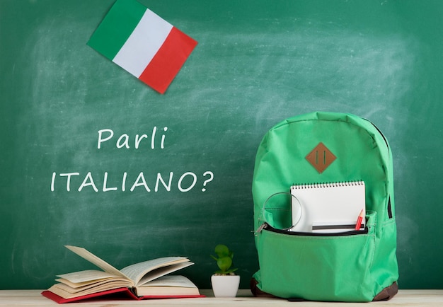 Concepto de aprendizaje de idiomas: mochila verde, pizarra con el texto "Parli italiano?", bandera de Italia, libros y cuadernos