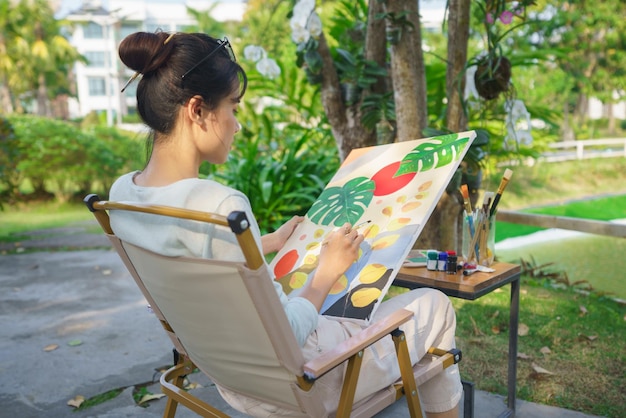 Concepto de artista Artista femenina sentada en el parque y pintando un cuadro sobre lienzo con felicidad