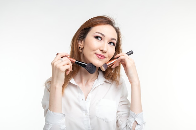 Concepto de artista, belleza y personas de maquillaje - hermosa joven coreana sosteniendo pinceles de maquillaje en la pared blanca