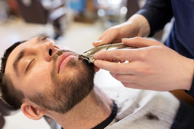 Concepto de aseo y personas: manos de hombre y barbero con recortadora o afeitadora cortando barba en la barbería