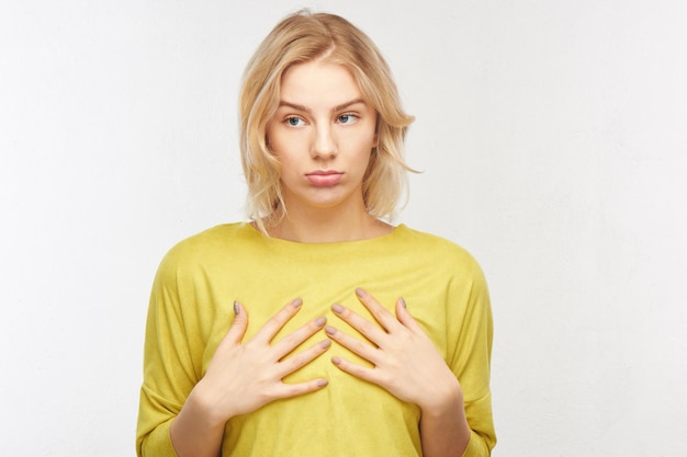 Concepto de aumento de senos, tamaño pequeño. Mujer joven molesta con una expresión facial disgustada toca sus pequeñas tetas en el estudio sobre un fondo blanco