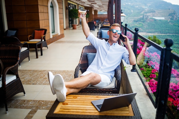 Foto concepto de autónomo. joven exitoso, empresario trabajando en una computadora portátil, sentado en la terraza tomando café y hablando por teléfono.