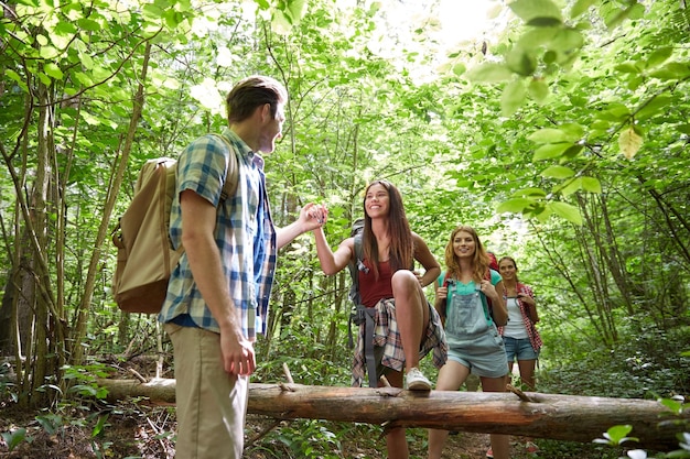 concepto de aventura, viajes, turismo, caminatas y personas - grupo de amigos sonrientes caminando con mochilas y trepando por el tronco de un árbol caído en el bosque