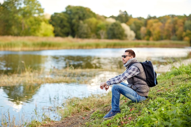 concepto de aventura, viajes, turismo, caminatas y personas - hombre con gafas de sol con mochila descansando en la orilla del río