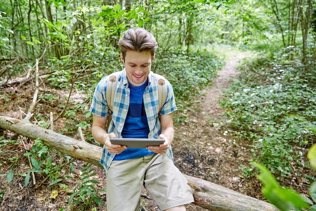 concepto de aventura, viajes, turismo, caminatas y personas - joven feliz con mochila y tablet pc sentado en un tronco de árbol caído en el bosque