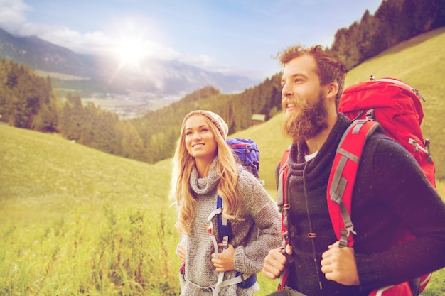 concepto de aventura, viajes, turismo, caminatas y personas - pareja sonriente caminando con mochilas sobre montañas alpinas y fondo de colinas