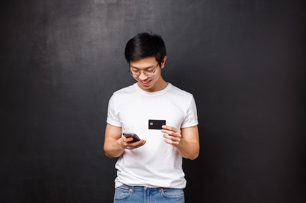 Concepto de banco, finanzas y pago. Joven asiático en camiseta, con tarjeta de crédito como insertar información de facturación en la aplicación de compras, mira la pantalla del teléfono móvil sonriendo, comprando ropa nueva en internet