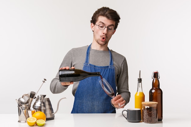 Concepto de barista, trabajador de café y barman. Retrato de hombre joven guapo tonto en delantal vertiendo vino en vaso y sonriendo divertido, necesita beber un sorbo de alcohol mientras prepara la cena,