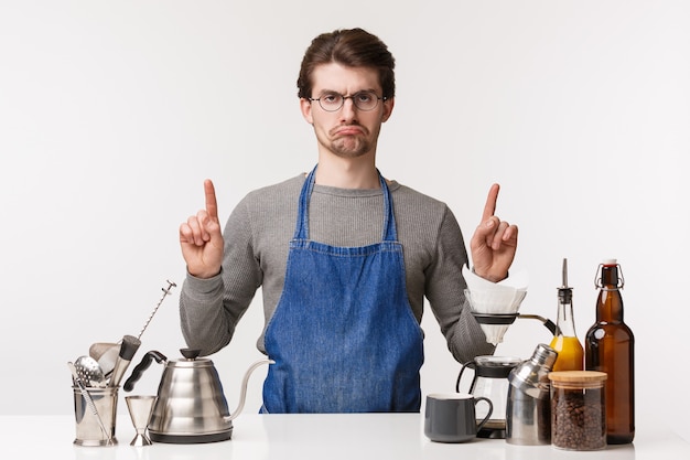 Concepto de barista, trabajador de café y barman. Retrato de un joven empleado enojado molesto y molesto que se siente insatisfecho y celoso apuntando hacia arriba mientras está de pie trabajando en la barra de bar, hacer café