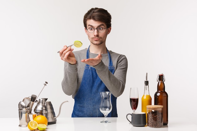 Concepto de barista, trabajador de café y barman. Retrato de joven varón caucásico en delantal con rodaja de limón, preparar un cóctel, haciendo una copa en el vaso, mirando centrado en la fruta,