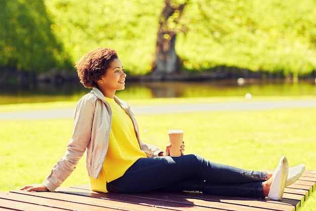 concepto de bebidas y personas - joven afroamericana sonriente o adolescente bebiendo café de una taza de papel en el parque de verano