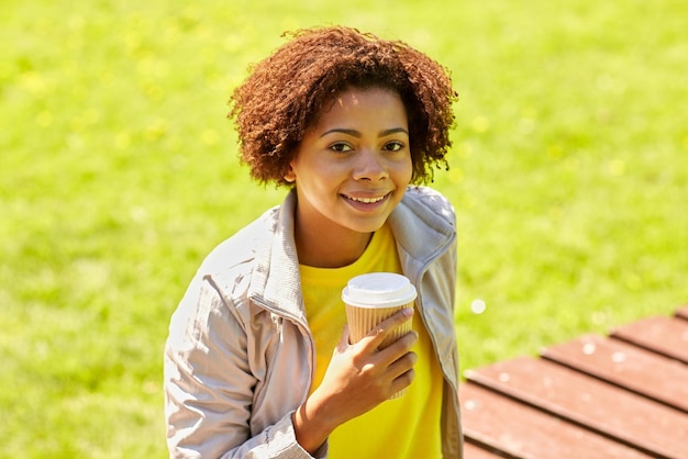 El concepto de bebidas y personas: una joven afroamericana sonriente o una adolescente bebiendo café de un vaso de papel al aire libre