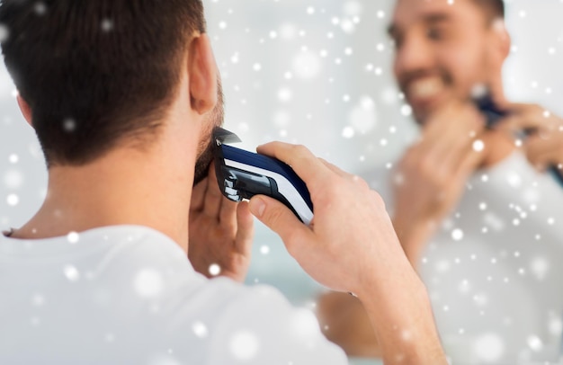 Concepto de belleza, afeitado, arreglo personal y personas: cierre de un joven que mira al espejo y se afeita la barba con un cortapelos o una afeitadora eléctrica en el baño de casa sobre la nieve