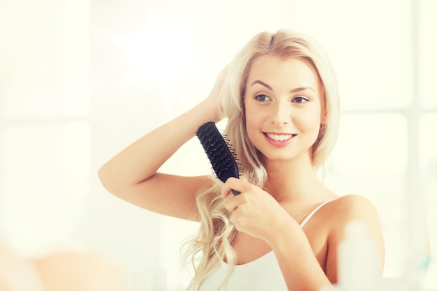 concepto de belleza, aseo y personas - mujer joven sonriente mirando al espejo y cepillando el cabello con peine en el baño de casa