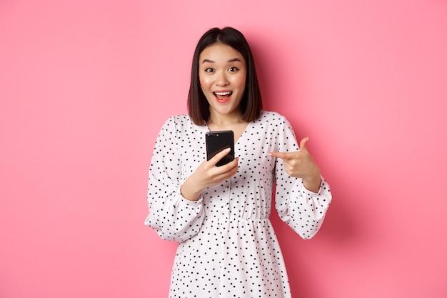 Concepto de belleza y compras en línea. Mujer asiática sorprendida y feliz apuntando al teléfono móvil, hablando de una oferta o aplicación promocional de Internet, de pie sobre rosa.