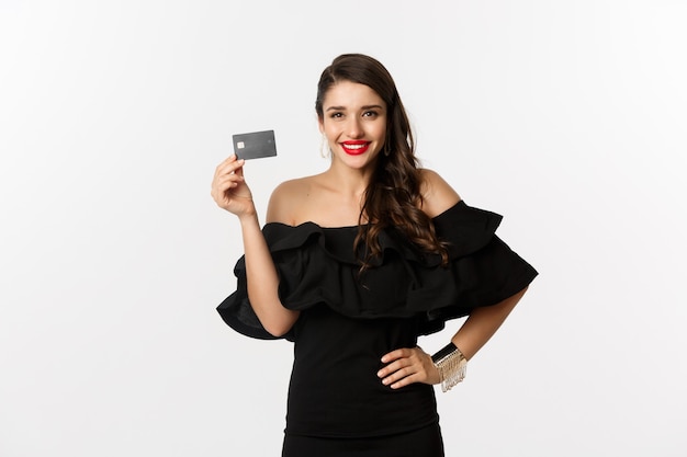Concepto de belleza y compras. Mujer bonita elegante con labios rojos, vestido negro, sonriendo feliz y mostrando tarjeta de crédito, de pie sobre fondo blanco.