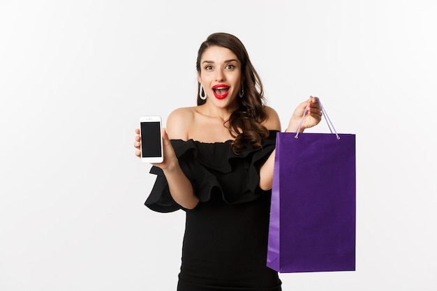 Concepto de belleza y compras. Mujer hermosa y elegante que muestra la pantalla del teléfono inteligente y el bolso, comprando en línea, de pie sobre fondo blanco.