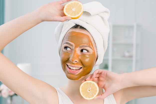 Concepto de belleza cuidado de la piel. La mujer caucásica atractiva en la toalla blanca con la máscara facial marrón en cara sostiene los agrios en su mano en espacio ligero. Procedimientos de spa y mascarilla en la piel.