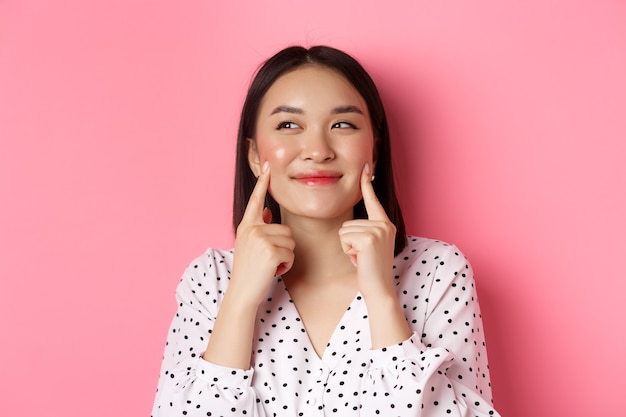 Concepto de belleza y cuidado de la piel. Primer plano de hermosa mujer asiática metiendo las mejillas y sonriendo satisfecho, de pie sobre fondo rosa