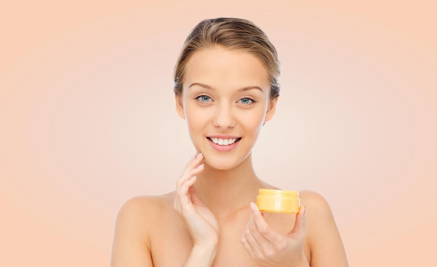 concepto de belleza, gente, cosméticos, cuidado de la piel y cosméticos - joven feliz aplicándose crema en la cara sobre fondo beige