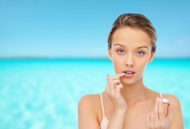 concepto de belleza, gente y cuidado de los labios - mujer joven que se aplica bálsamo labial en los labios sobre el fondo azul del mar y el cielo