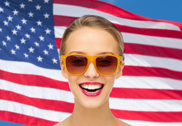 concepto de belleza, gente, nacionalidad y patriotismo - mujer joven sonriente con gafas de sol con lápiz labial rosa en los labios sobre el fondo de la bandera americana