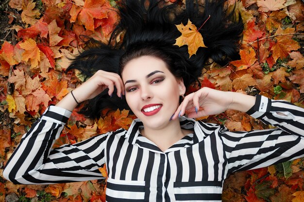 Concepto de belleza, gente, temporada y salud - chica guapa está acostada en hojas de otoño rojas amarillas