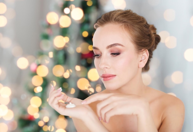 concepto de belleza, gente, vacaciones, cuidado de la piel y cosméticos - mujer joven con crema hidratante a mano sobre fondo de luces de árbol de Navidad