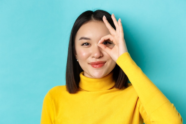 Concepto de belleza y maquillaje. Cerca de una chica asiática despreocupada que muestra el signo de OK en los ojos y sonriendo, mirando feliz a la cámara, de pie sobre fondo azul.