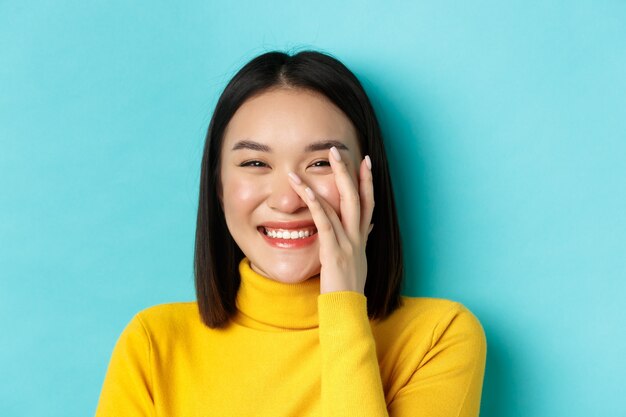 Concepto de belleza y maquillaje. Cerca de la hermosa mujer asiática ruborizándose y riendo, tocando la piel sana brillante, sonriendo feliz, de pie sobre fondo azul.