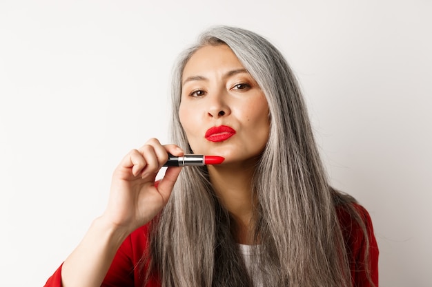 Concepto de belleza y maquillaje. Labios fruncidos de la mujer mayor asiática hermosa, mostrando el lápiz labial rojo y mirando descarado a la cámara, fondo blanco.