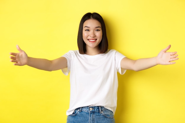 Concepto de belleza y moda. Amable mujer asiática extendió las manos y sonriendo, esperando abrazos, invitándote, dando la bienvenida a alguien y luciendo feliz, de pie sobre un fondo amarillo.