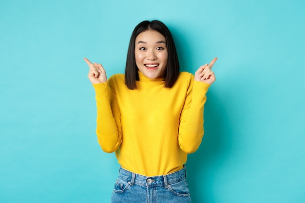 Concepto de belleza y moda. Chica asiática alegre mostrando dos ofertas promocionales, señalando con el dedo hacia los lados en los anuncios de izquierda y derecha y sonriendo, de pie sobre fondo azul.