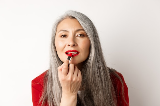 Concepto de belleza y moda. Elegante mujer madura asiática con cabello gris, mirando en el espejo y aplicar lápiz labial rojo, de pie sobre fondo blanco.