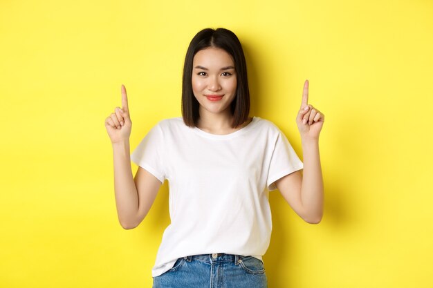 Concepto de belleza y moda. Hermosa mujer asiática en camiseta blanca apuntando con el dedo hacia arriba, de pie sobre fondo amarillo.