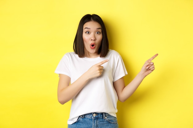 Concepto de belleza y moda. Hermosa mujer asiática en camiseta blanca apuntando con el dedo hacia la derecha, demuestra el logotipo de pie sobre fondo amarillo