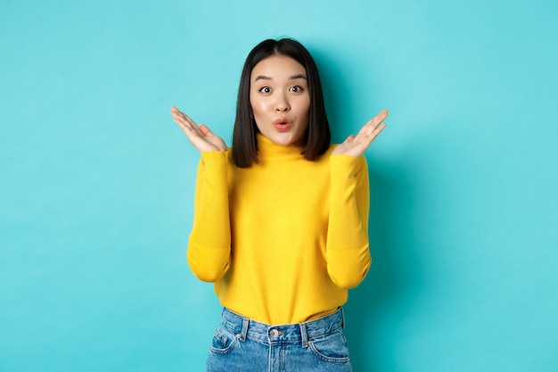 Concepto de belleza y moda. Imagen de una niña japonesa emocionada y sorprendida diciendo wow con asombro, levantando las manos cerca de la cara, de pie contra el fondo azul.