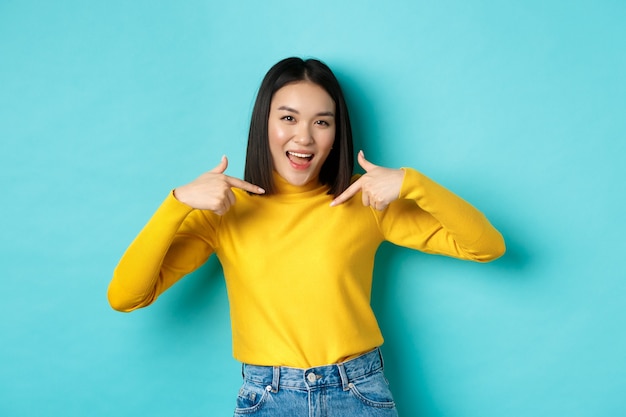 Concepto de belleza y moda. Mujer asiática segura y atrevida apuntando a sí misma, sonriendo descaradamente a la cámara, siendo profesional, de pie sobre fondo azul.